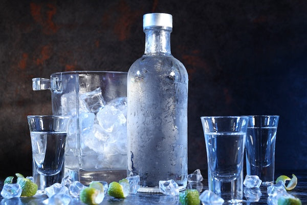 A frosted bottle of clear liquor stands next to a glass ice bucket filled with ice cubes. Two shot glasses are placed beside the bottle, and scattered ice cubes and lime wedges surround them on a dark surface.