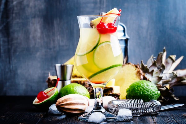 A refreshing tropical drink is served in a tall glass, garnished with pineapple slices, lime wheels, and a cherry. Ice cubes surround the drink on a dark wooden surface. Pineapples, limes, a cocktail shaker, and a strainer are in the background.