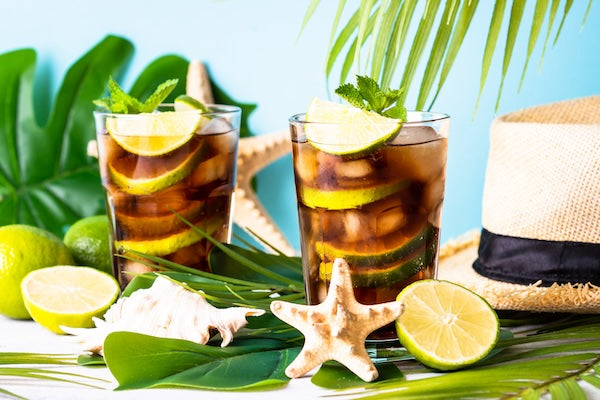 Two glasses of iced drinks with lime slices and mint garnishes, surrounded by tropical leaves, starfish, seashells, whole and halved limes. A straw hat partially visible on the right. The background is a vibrant light blue, evoking a summer beach vibe.