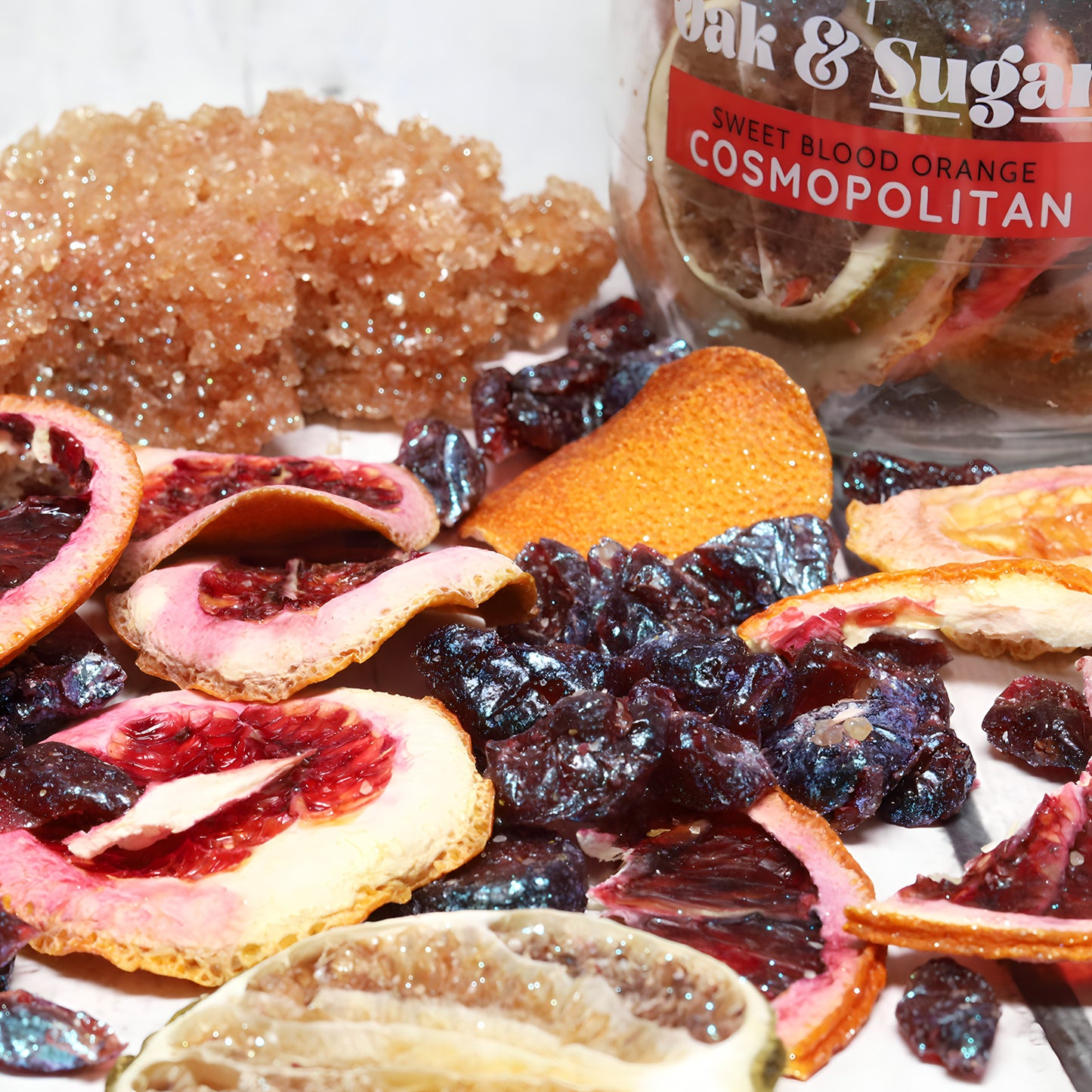 A display of dried citrus fruits, including blood oranges and lime, along with dried cranberries. A jar labeled "Oak & Sugar Sweet Blood Orange Cosmopolitan Cocktail Infusion Kit" partially filled with dried fruit slices and sugar is in the background, adorned with drinkable food-safe glitter for added sparkle.