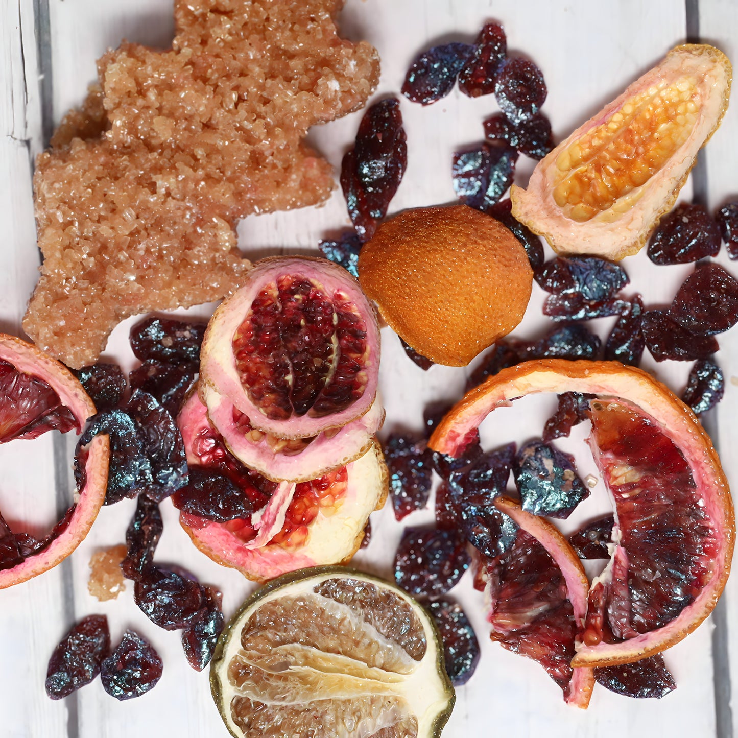 An assortment of dried fruits and crystals is spread out on a white wooden surface. The arrangement includes slices of dried citrus fruits, a shriveled orange segment, and clusters of dark, shiny dried berries or cherries, along with a large handcrafted signature Oak & Sugar Sweet Blood Orange Cosmopolitan Cocktail Infusion Kit.