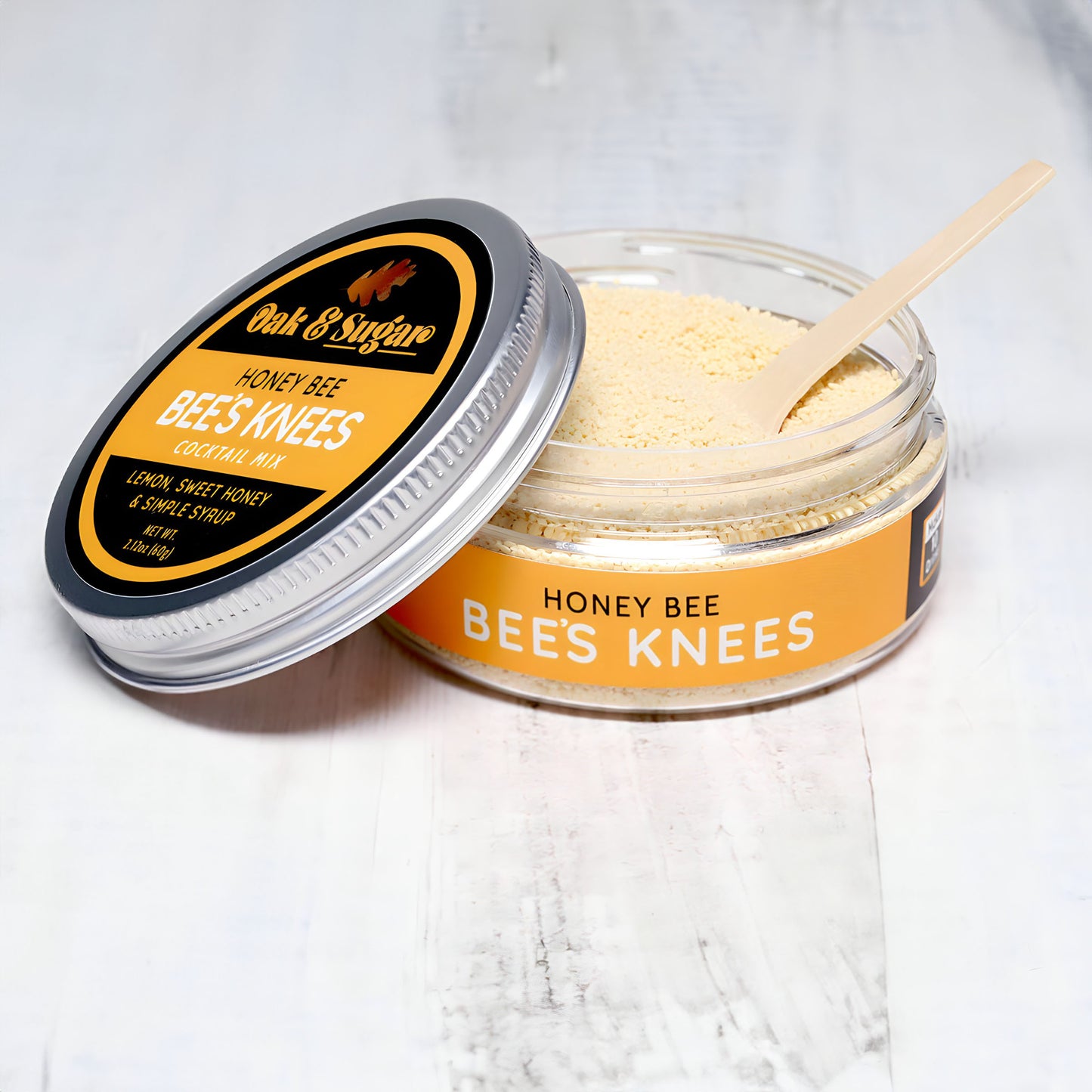 A jar of Oak & Sugar’s Honey Bee Bees Knees Cocktail Mixer with a silver lid partially removed, revealing a powdery mixture inside. A small wooden spoon rests in the jar. The jar is placed on a light-colored wooden surface.