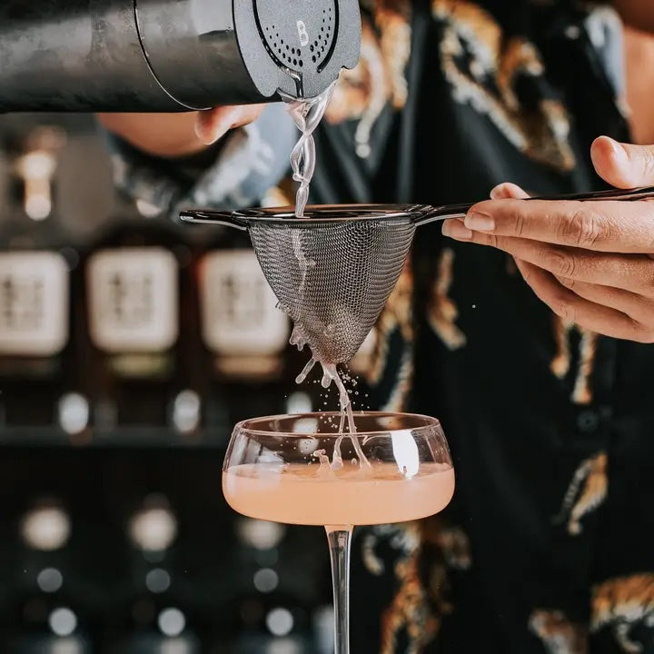 A person pours a cocktail through a Bull in China Fine Mesh Cocktail Strainer, Large into a coupe glass. The liquid streams from a cocktail shaker, with bottles visible in the blurry background. The person is wearing a dark, patterned shirt.