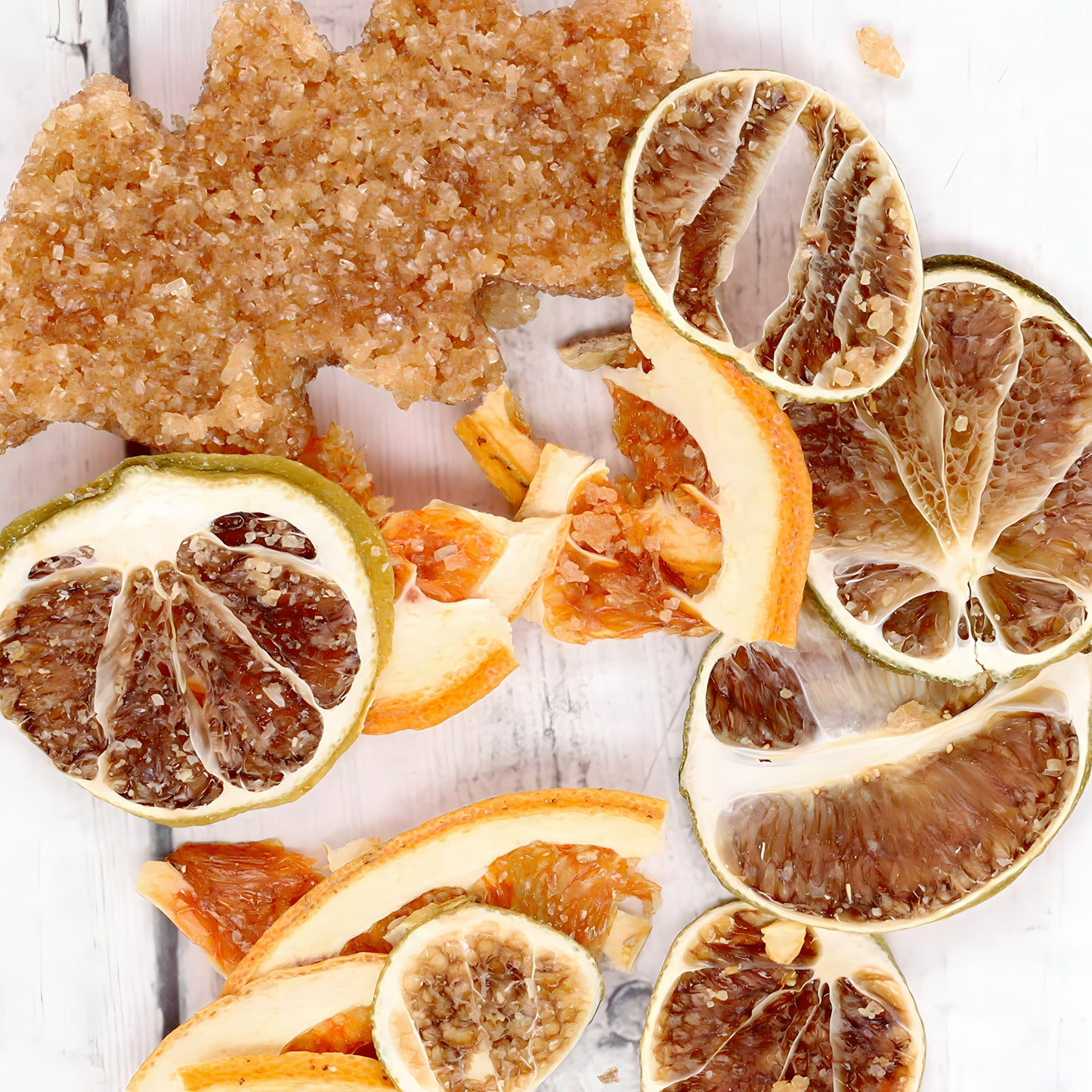 A variety of dried fruits and a cluster of brown rock sugar crystals on a light wooden surface. The dried fruits, perfect for the Oak & Sugar The Denton Grapefruit Moscow Mule Cocktail Infusion Kit, include slices of oranges, lemons, and limes, all showcasing a textured, slightly curled appearance.