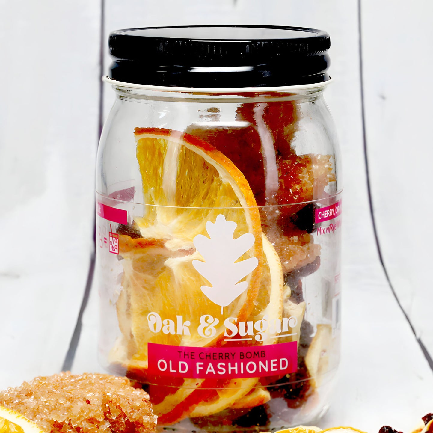 A clear glass jar with a black lid labeled "Oak & Sugar" contains ingredients for The Cherry Bomb Old Fashioned Infusion Kit. Inside, you'll find dried orange slices and sugared candies. Perfect for enhancing your cocktail experience, try it with Angostura bitters. The label features a white oak leaf logo and red text on a white background.