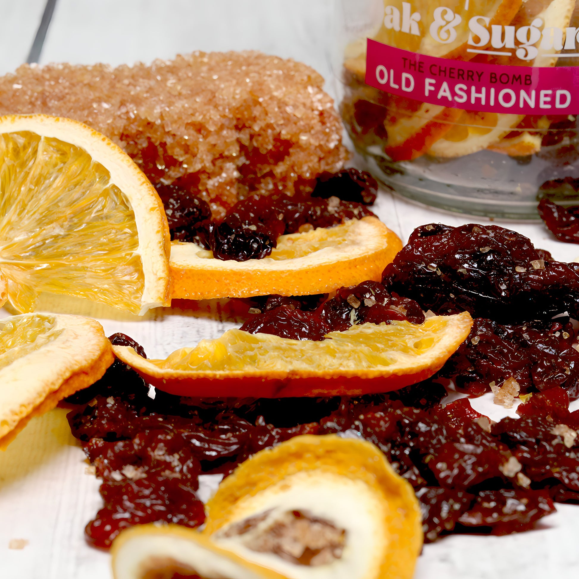 A close-up of dried orange slices, dark dried cherries, and a piece of brown sugar on a white wooden surface. In the background, there is a partially visible jar labeled "The Cherry Bomb Old Fashioned Infusion Kit" by Oak & Sugar, hinting at the perfect cocktail with Angostura bitters waiting inside.