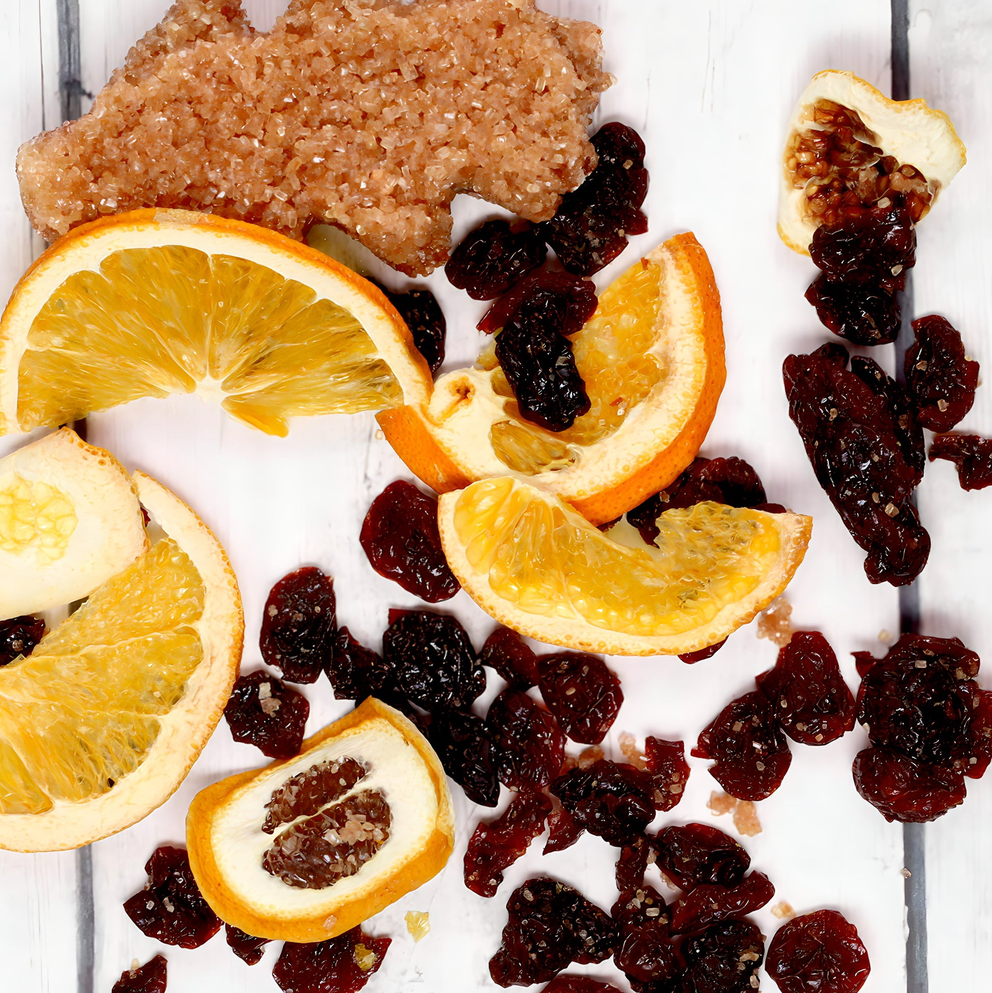 A flat lay on a white wooden surface features dehydrated orange slices, assorted dried fruits including raisins and fig slices, and a brown cluster of granulated sugar. Scattered randomly to create a rustic, natural look, these ingredients could be perfect for your next cocktail using The Cherry Bomb Old Fashioned Infusion Kit by Oak & Sugar.