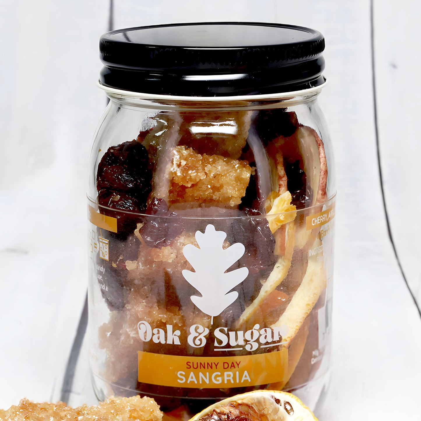 A clear mason jar with a black lid is filled with dried fruits and sugar crystals. The label reads "Oak & Sugar" featuring a white oak leaf logo and "Sunny Day Sangria Infusion Kit." This fruity blend sits on a white wooden surface with a partially visible sliced orange in the foreground.