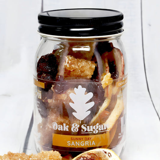 A clear mason jar with a black lid is filled with dried fruits and sugar crystals. The label reads "Oak & Sugar" featuring a white oak leaf logo and "Sunny Day Sangria Infusion Kit." This fruity blend sits on a white wooden surface with a partially visible sliced orange in the foreground.