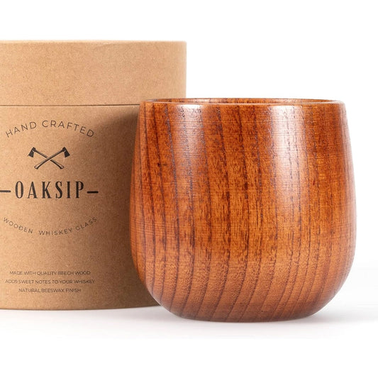 A Finished Beech Wood Bourbon/Whiskey Drinking Glass is positioned next to a cylindrical cardboard container. The container, labeled "Oaksip Finished Beech Wood Bourbon/Whiskey Drinking Glass," features small crossed ax illustrations. The glass has smooth, polished wood with visible grain patterns, showcasing its unique design.