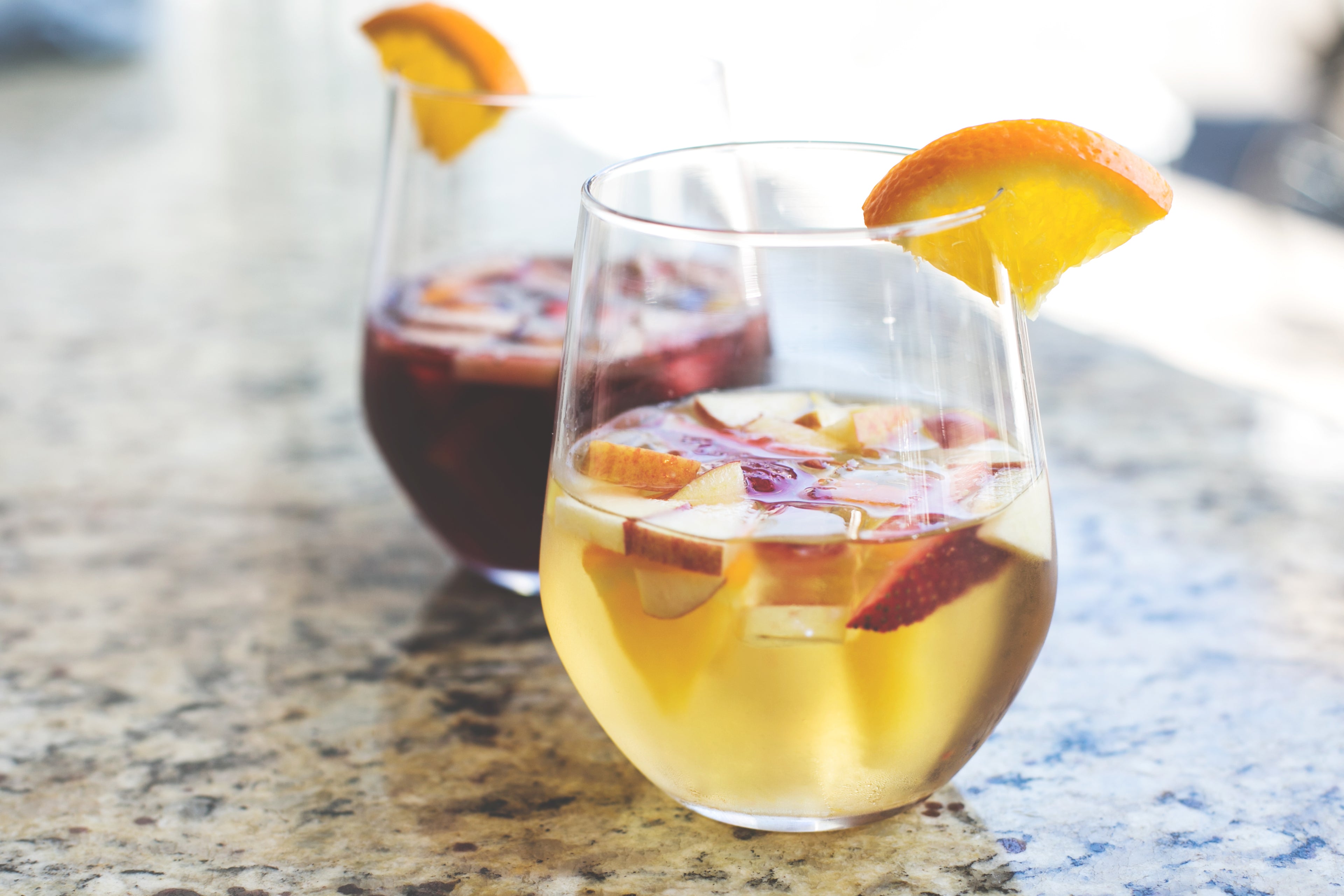 Two glasses of sangria on a granite countertop, one containing a refreshing white sangria with fruit slices and an orange wedge, and the other a vibrant red sangria, both garnished with an orange wedge. The glasses are filled with ice and look perfect for a sunny day.