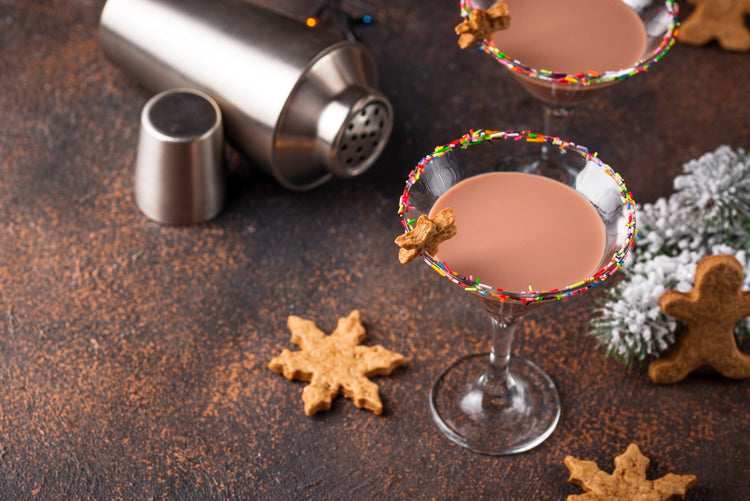 Two martini glasses filled with a creamy cocktail, adorned with a sprinkles rim and a gingerbread cookie. A shaker and jigger are in the background, alongside snowflake and gingerbread cookies on a rustic surface.