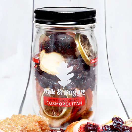 A jar labeled "Oak & Sugar Sweet Blood Orange Cosmopolitan Cocktail Infusion Kit," containing dried fruits and citrus slices for a delightful cocktail infusion. The jar with a black lid is set on a white rustic surface with additional dried fruit, coarse sugar, and an artisan oak leaf sugar cube scattered around.