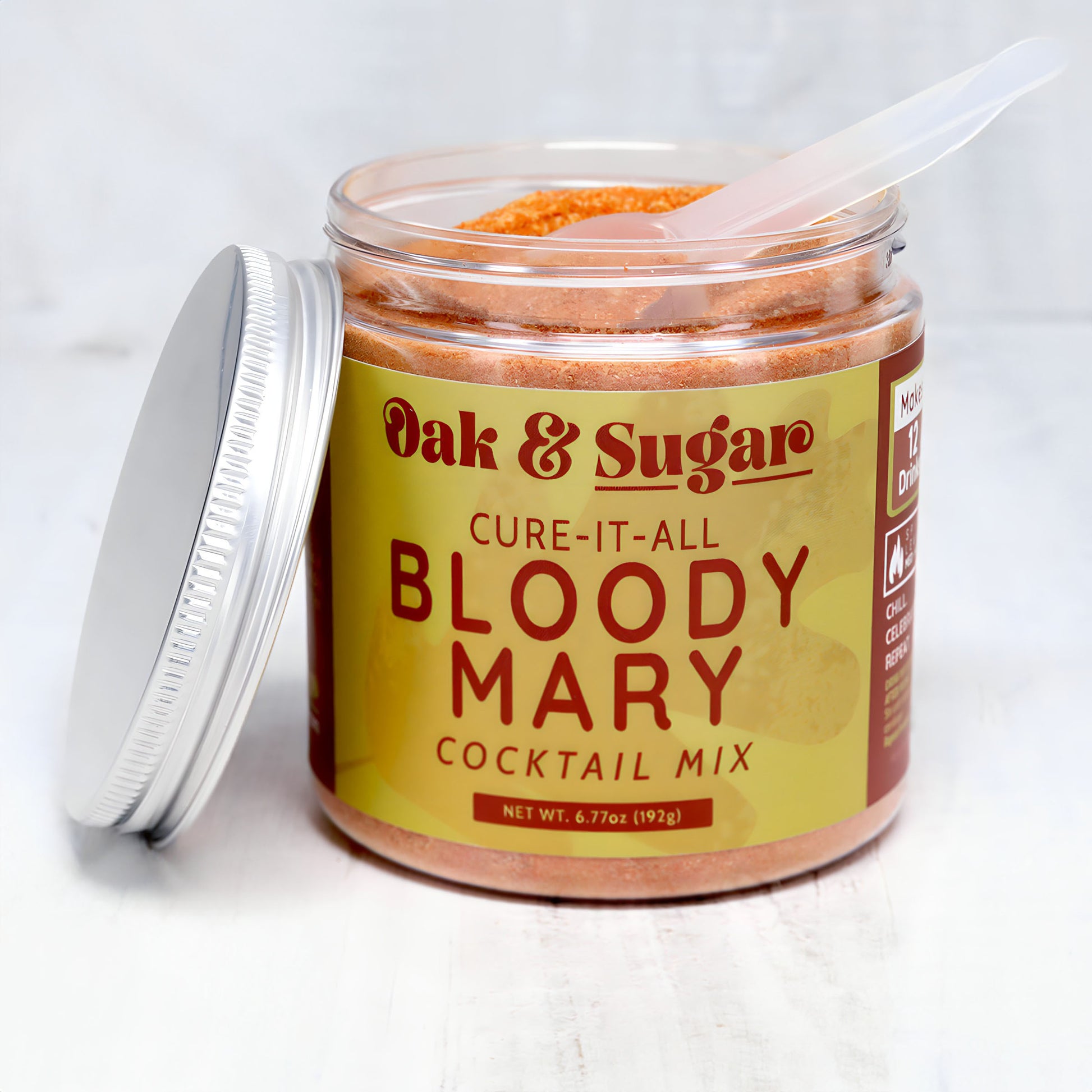 A jar of Oak & Sugar brand "Cure-It-All Bloody Mary Cocktail Mix" on a pale surface. The jar is open with the lid placed beside it and a small, clear plastic scoop sticking out of the mix. The mix, perfect for brunch cocktails, appears to be a coarse, orange-colored powder.