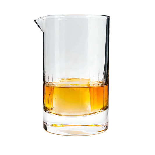 A premium high-quality Premium Cocktail Mixing Glass, 20oz by Thousand Oaks Barrel Co. filled partially with amber-colored liquid, featuring a single large ice cube. The beaker has a small spout, and its surface is slightly frosted near the bottom. The background is plain white.