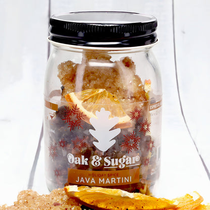 Clear glass jar with a black lid, labeled "Oak & Sugar Holiday Spice Java Martini Cocktail Infusion Kit - LIMITED EDITION." The jar is filled with brown sugar crystals, and dried orange slices are visible inside. Additional brown sugar crystals and dried orange slices are arranged outside the jar.