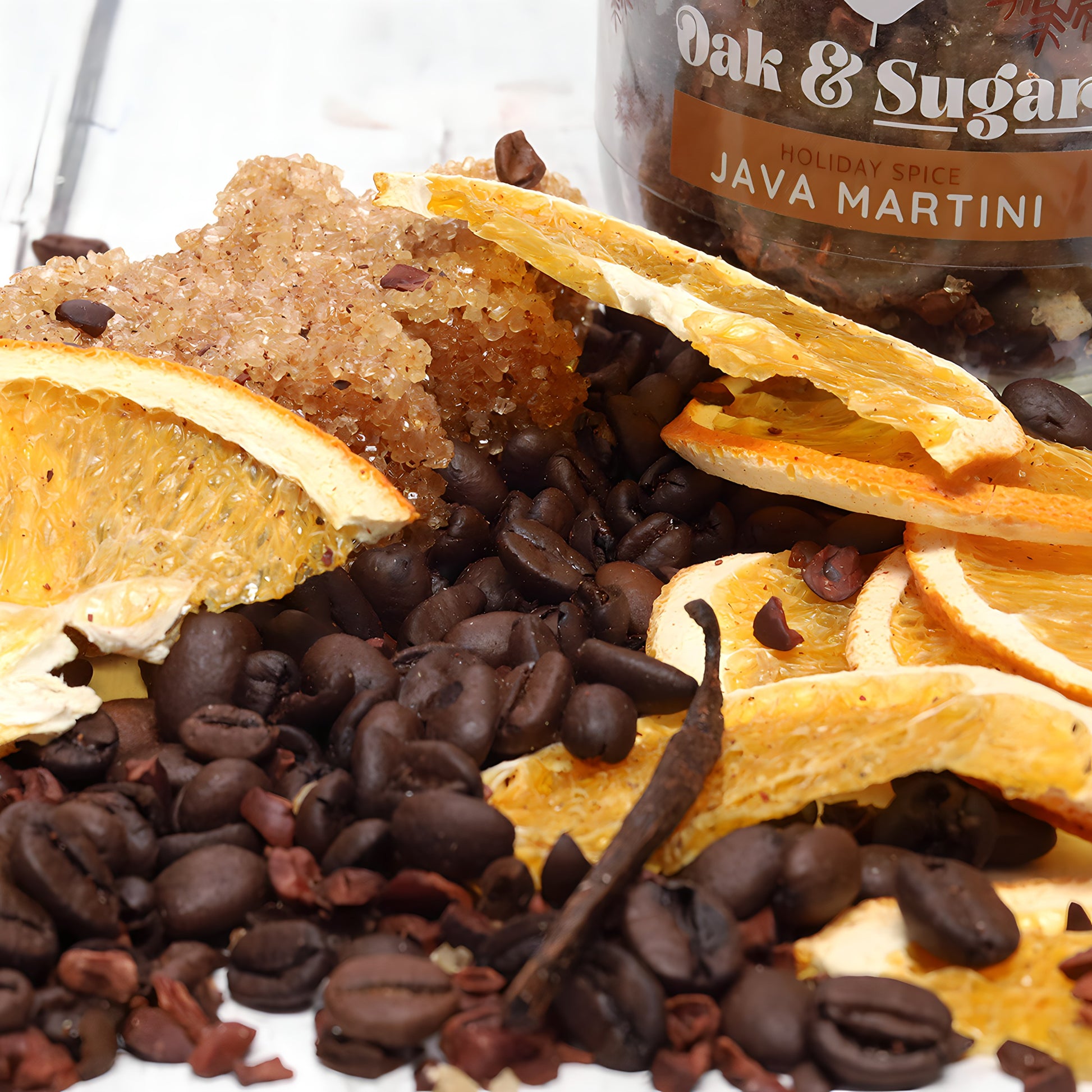 A close-up of a holiday spice mix for a Spiced Espresso Martini. The mix includes dried orange slices, vanilla beans, coffee beans, cocoa nibs, and coarse brown sugar. A jar labeled "Oak & Sugar Holiday Spice Java Martini Cocktail Infusion Kit - LIMITED EDITION" is partially visible in the background.