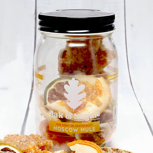A clear jar labeled "Oak & Sugar: The Denton Grapefruit Moscow Mule Cocktail Infusion Kit" contains dried grapefruit slices and other garnishes, perfect for an infusion kit. The jar has a black lid and sits on a white surface adorned with more dried citrus flavors.