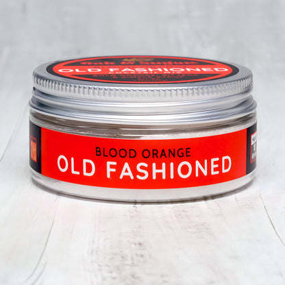 A round metal tin with a silver lid, labeled "Blood Orange Old Fashioned" in bold red letters, is placed against a light wood background. This versatile tin of Blood Orange Old Fashioned Cocktail Mix by Oak & Sugar is perfect for storing cocktail mix or spices.