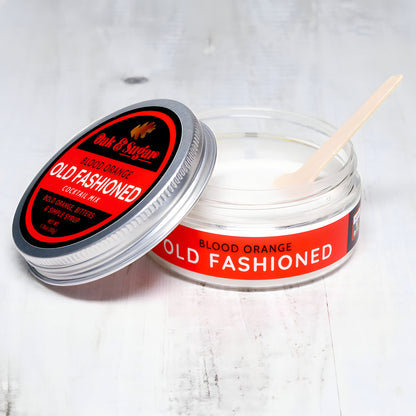 A clear jar of "Blood Orange Old Fashioned Cocktail Mix" with a red label is open, revealing a white powder inside. The jar's lid rests beside it, showcasing the "Oak & Sugar" logo and name. A small wooden spoon is inside the jar on a white surface, ready to blend the bitters into your cocktail mix.
