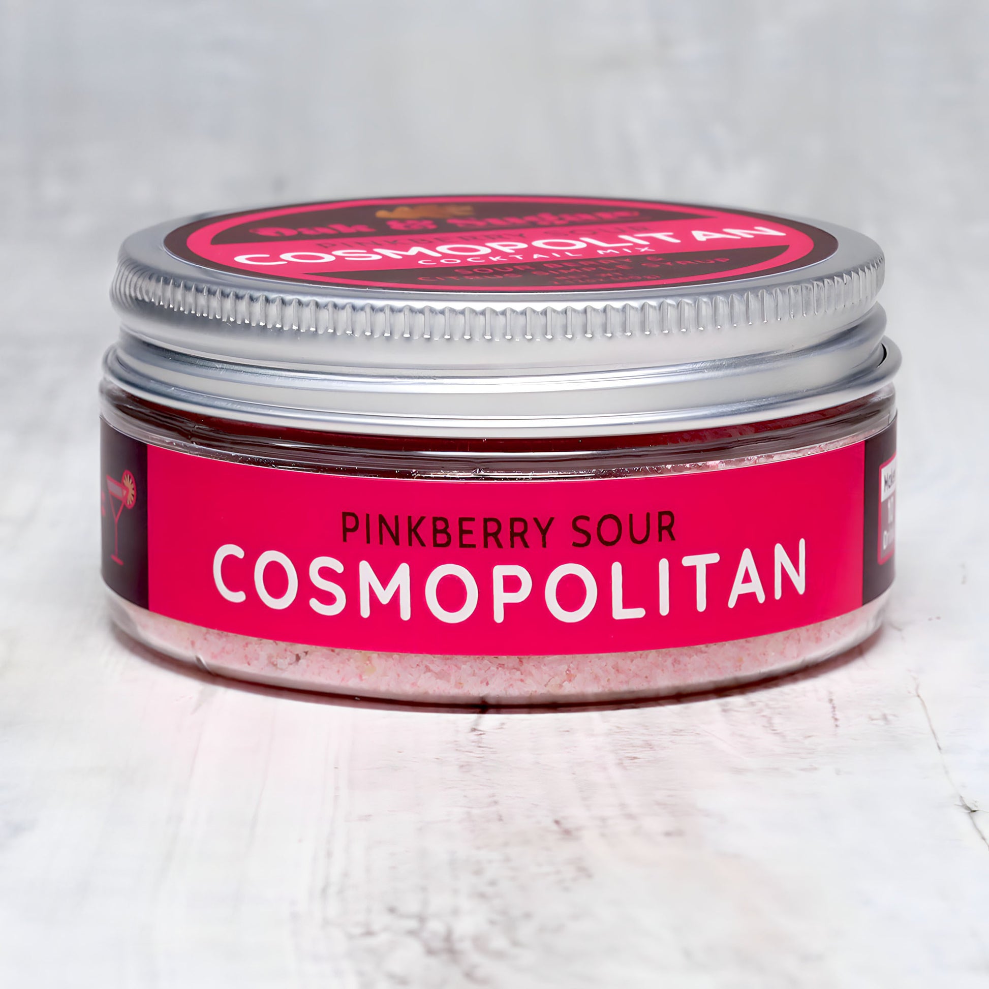 A small, round jar with a silver lid labeled "Pinkberry Sour Cosmopolitan Cocktail Mix" by Oak & Sugar. The label is primarily pink and red, with white text. The jar sits on a light, textured surface, and a portion of pink powder is visible inside. This delightful blend brings to mind the iconic posts from *Sex and the City*.
