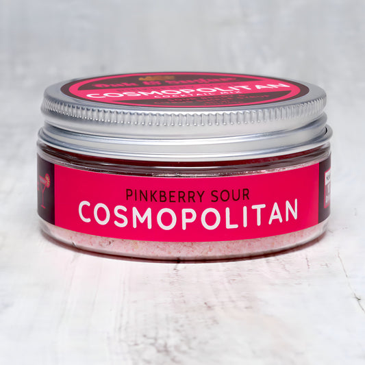 A small, round jar with a silver lid labeled "Pinkberry Sour Cosmopolitan Cocktail Mix" by Oak & Sugar. The label is primarily pink and red, with white text. The jar sits on a light, textured surface, and a portion of pink powder is visible inside. This delightful blend brings to mind the iconic posts from *Sex and the City*.