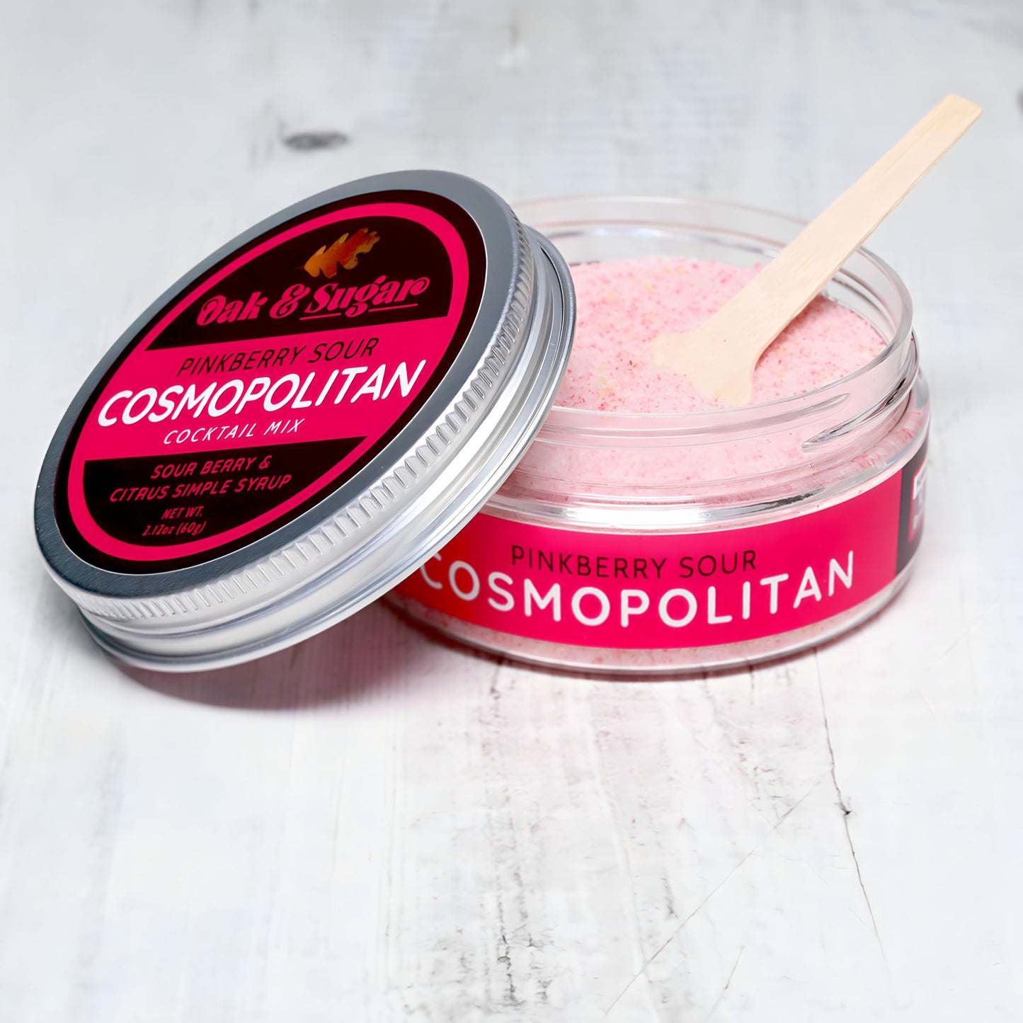 A glass jar of Oak & Sugar Pinkberry Sour Cosmopolitan Cocktail Mix is displayed on a light-colored surface. The jar is open, revealing pink powder inside, with a small wooden scoop resting on the edge. The label is red with white and pink text, reminiscent of a chic *Sex and the City* vodka cocktail moment.