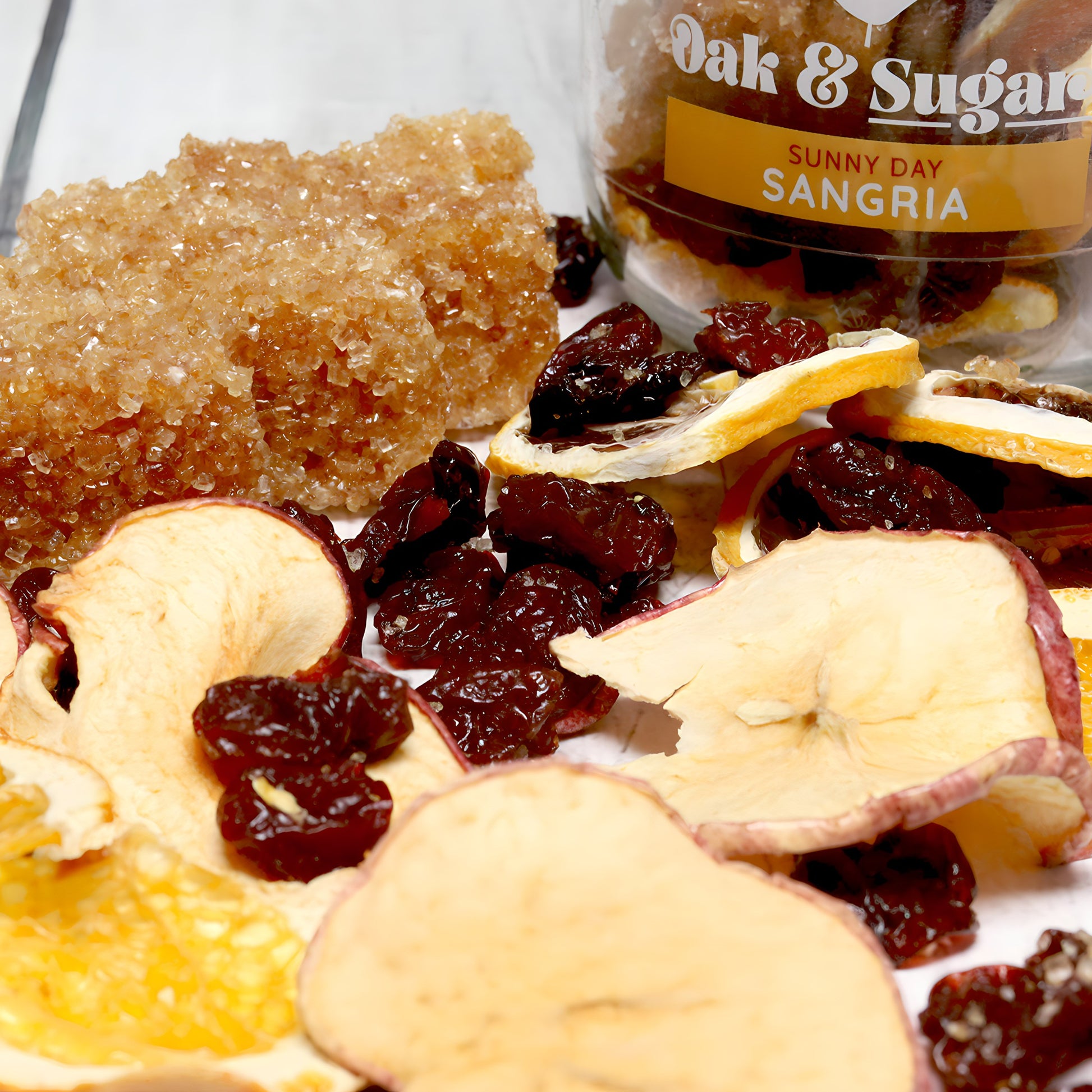 Close-up of dried apple slices, cranberries, and other mixed dried fruits scattered on a surface. A chunk of golden-brown rock sugar sits at the back. On the right, a clear plastic container labeled "Oak & Sugar: Sunny Day Sangria Cocktail Infusion Kit" is partially visible, showcasing a delightful fruity blend.