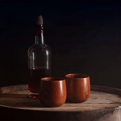 A bottle with a cork stopper filled partially with dark liquid stands on an aged wooden barrel. In front of the bottle are two Oaksip Finished Beech Wood Bourbon/Whiskey Drinking Glasses. The background is dark, highlighting the rustic and cozy ambiance o